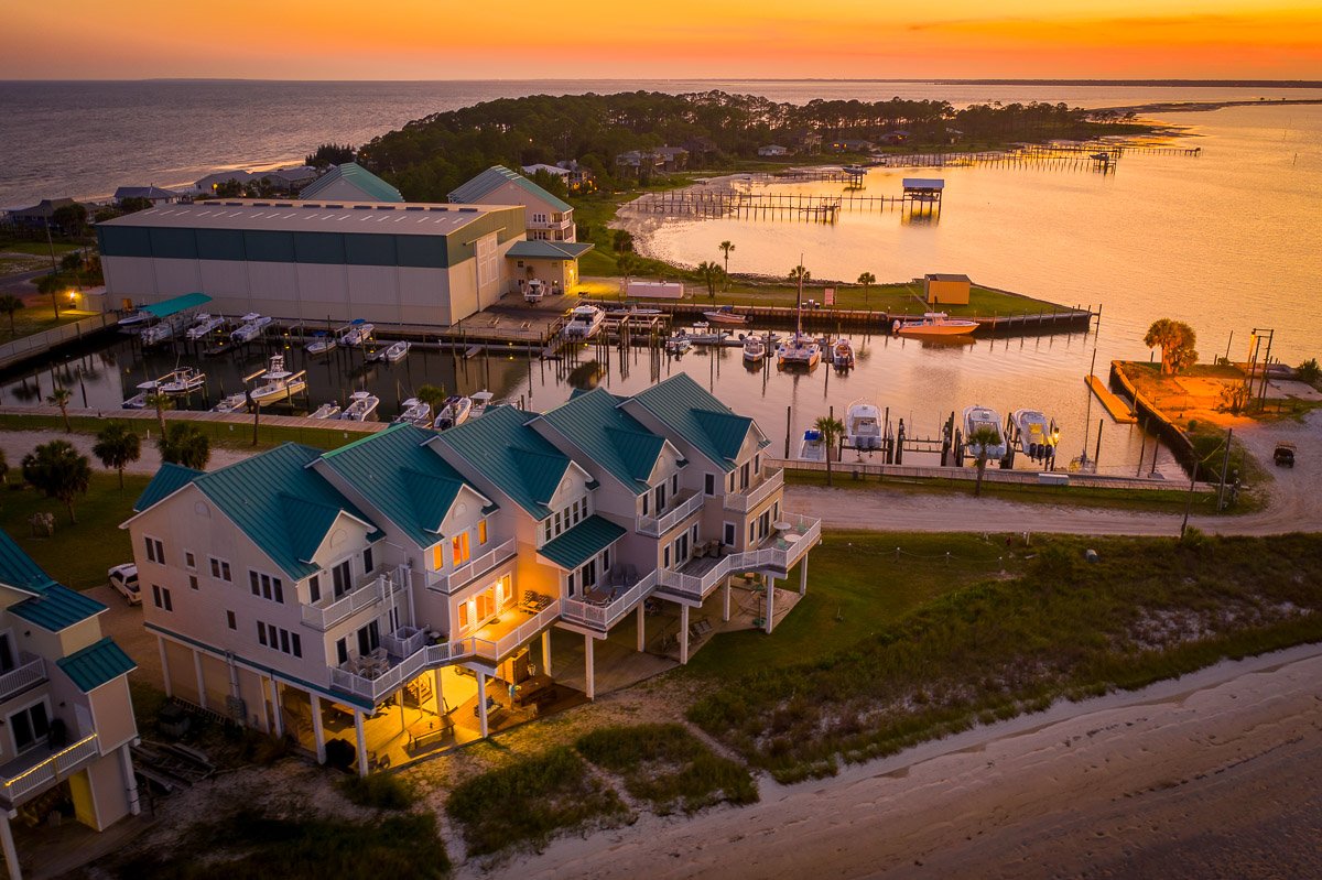 Professional aerial real estate photography of luxury waterfront homes beside a marina at sunset, with boats, docks, and expansive coastal views.