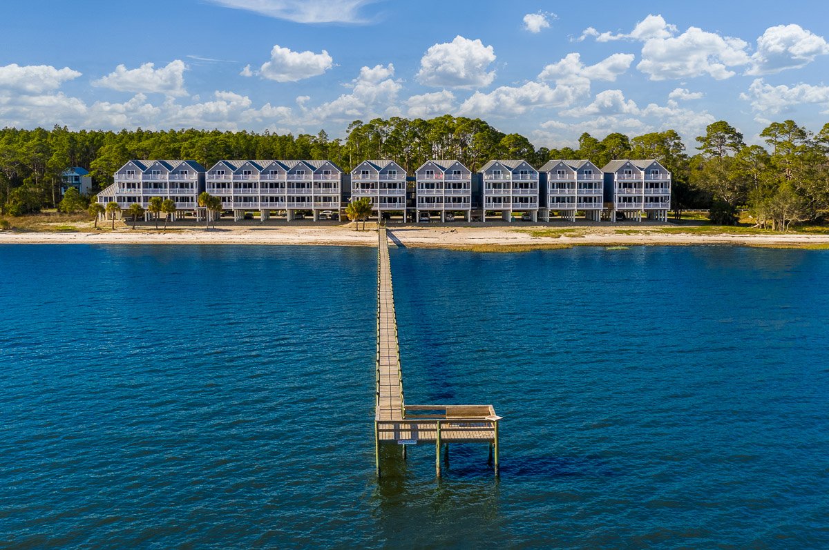 Professional aerial real estate photography of waterfront townhomes with a private pier, blue water, sandy shoreline, and expansive coastal views.