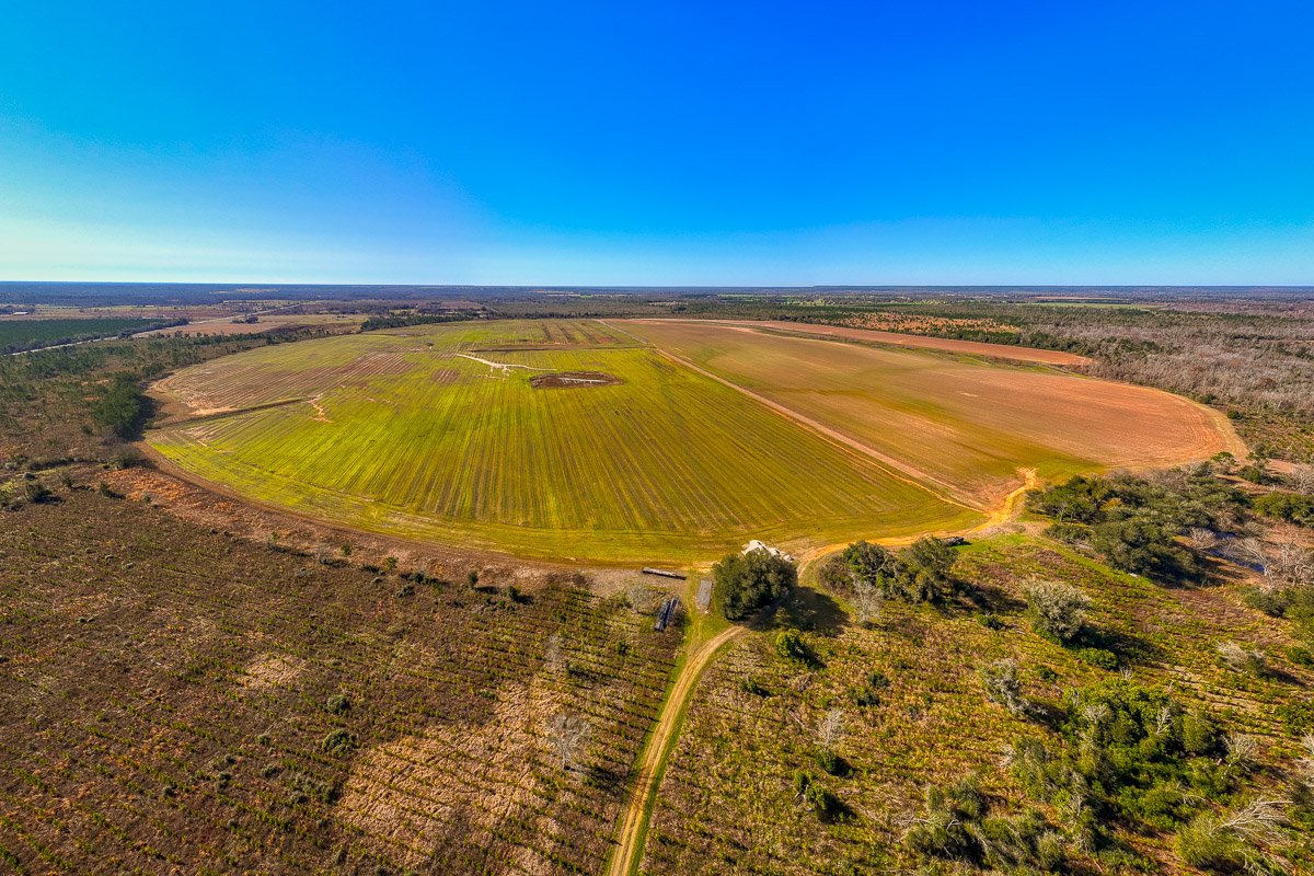 Professional aerial real estate photography of more than 100 acres of farmland, showcasing expansive open fields, rural access roads, and sweeping countryside views.