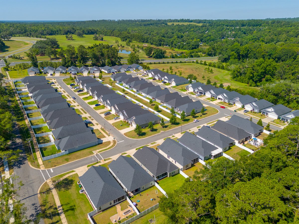 Professional aerial real estate photography of a large residential community with rows of modern homes, neighborhood streets, green space, and surrounding wooded landscape.