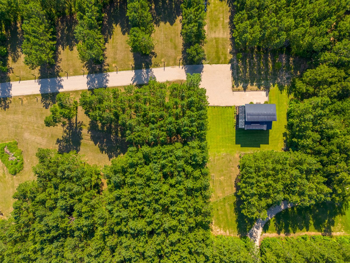 Professional aerial real estate photography of a luxury new construction home on a large private lot, shown from a top-down drone view with surrounding trees and expansive grounds.