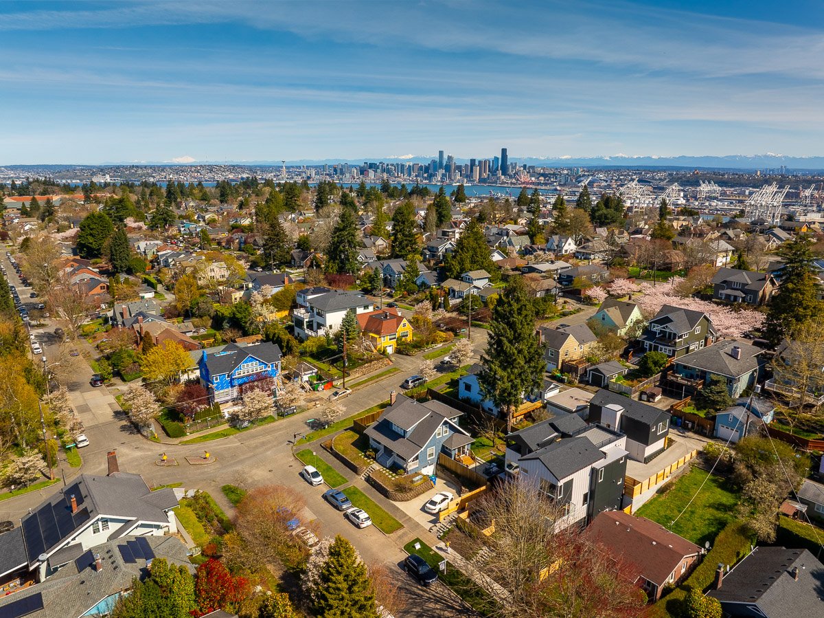 Professional aerial real estate photography of a Seattle residential neighborhood with the downtown skyline, Port of Seattle, and mountain views in the distance.