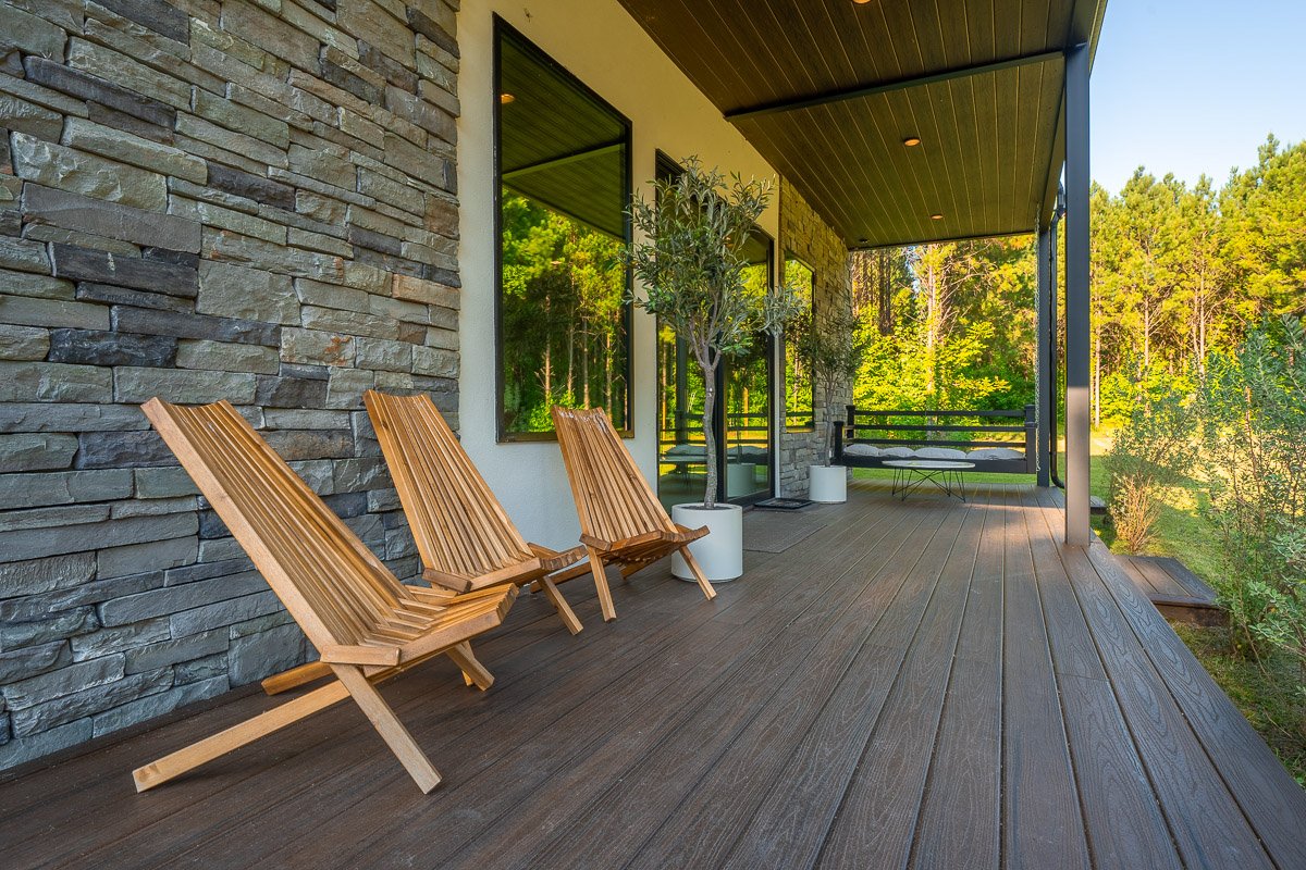 Professional real estate photography of a luxury modern home’s covered outdoor living space with stone walls, wood decking, contemporary seating, and a private wooded setting.