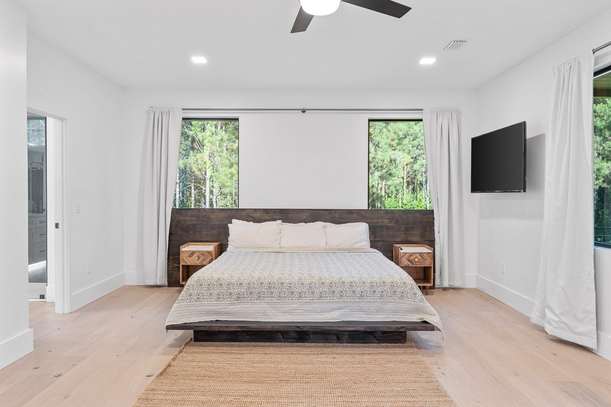 Professional real estate photography of a modern Seattle primary bedroom with large windows, minimalist furnishings, light wood floors, and peaceful wooded views.