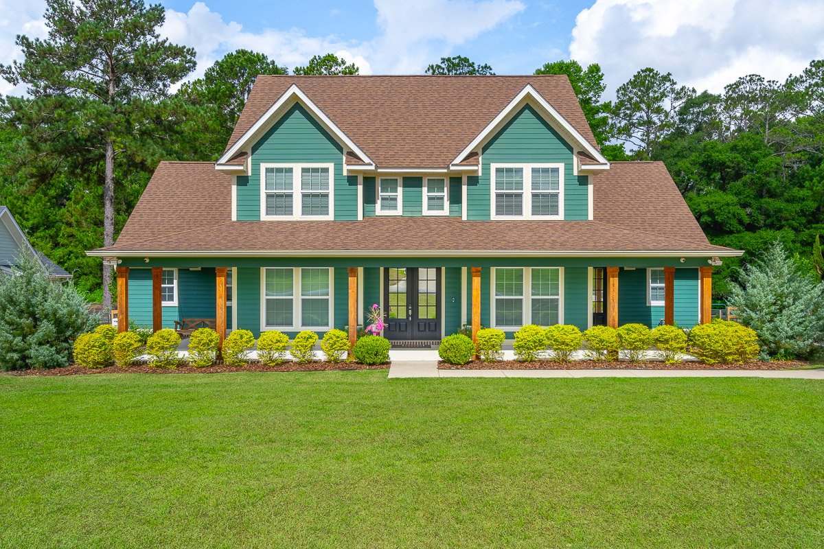 Professional real estate exterior photography of a large two-story home with green siding, covered front porch, manicured landscaping, and strong curb appeal.