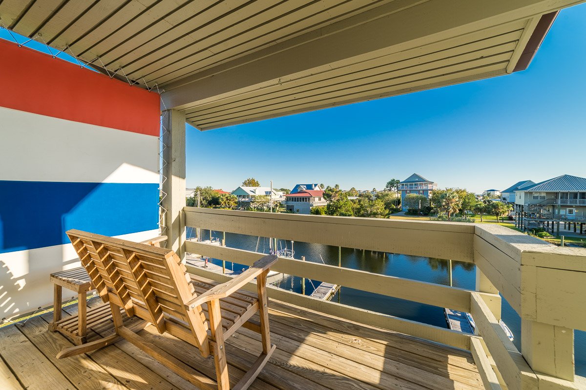 Professional real estate photography of a waterfront balcony overlooking a marina and coastal homes, with wooden seating and expansive water views.