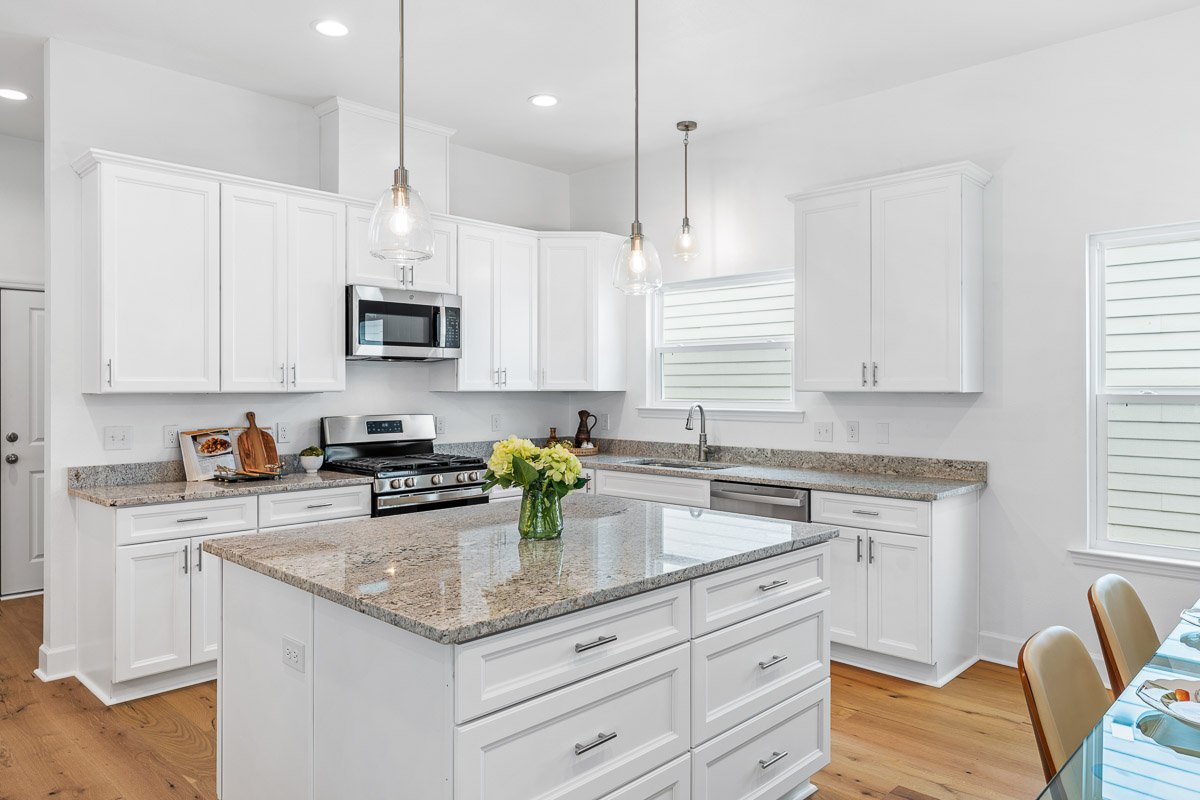Professional real estate photography of a bright Seattle kitchen with white cabinetry, large granite island, stainless steel appliances, pendant lighting, and hardwood floors.