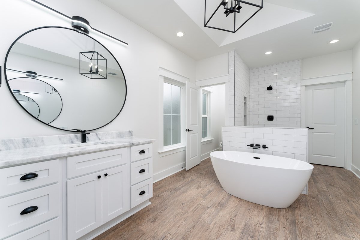 Luxury bathroom real estate photo captured with flambient lighting, featuring a freestanding soaking tub, large round mirror, marble vanity, walk-in shower, and bright modern finishes.