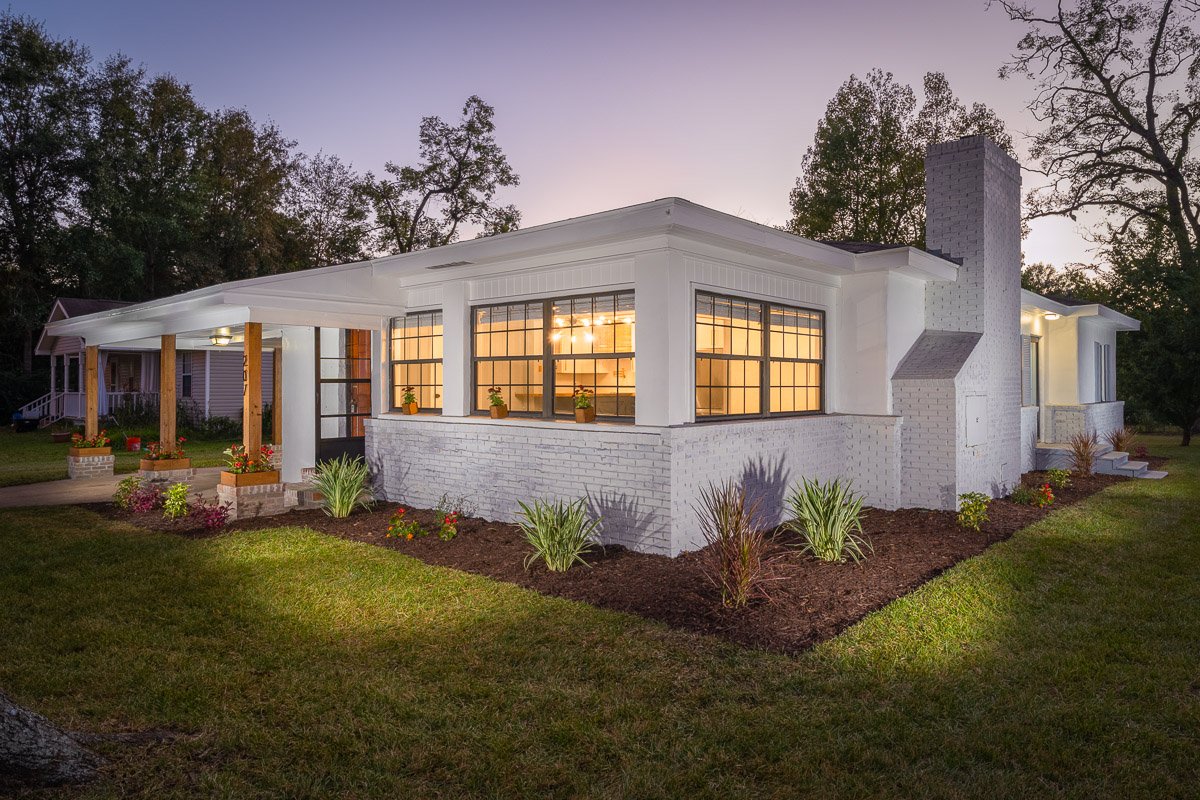 Professional twilight real estate photography of a renovated white brick home with warm interior lighting, landscaped yard, covered porch, and inviting curb appeal.