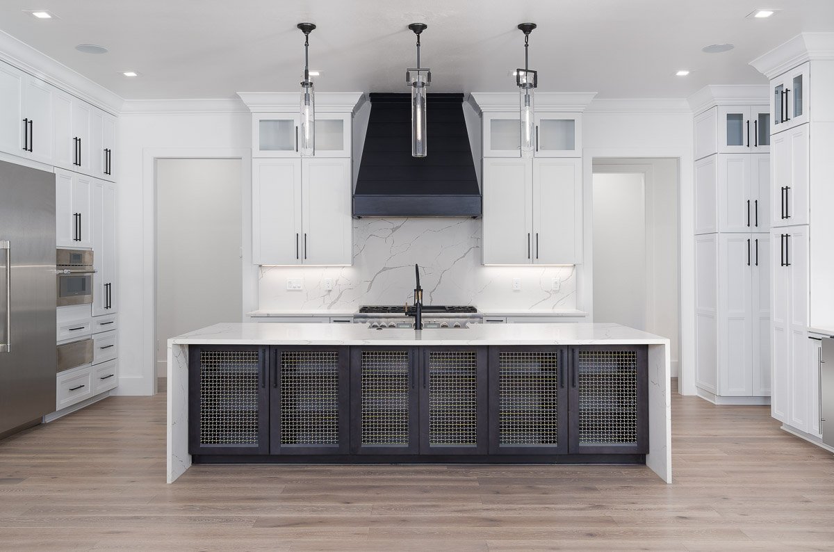 Flambient real estate kitchen photography of a bright luxury kitchen with a large waterfall island, black range hood, white shaker cabinetry, quartz backsplash, and modern pendant lighting.