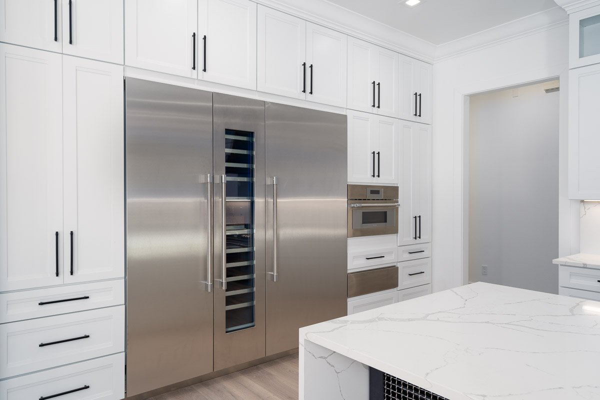 Professional flambient real estate photography of a luxury kitchen featuring custom white cabinetry, a built-in stainless steel refrigerator and wine column, integrated wall ovens, and a large quartz waterfall island.