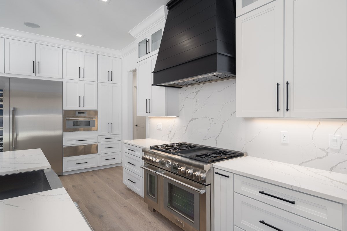 Professional real estate photography of a luxury Seattle kitchen with white shaker cabinetry, quartz countertops and backsplash, a commercial-style gas range, and a dramatic black range hood.