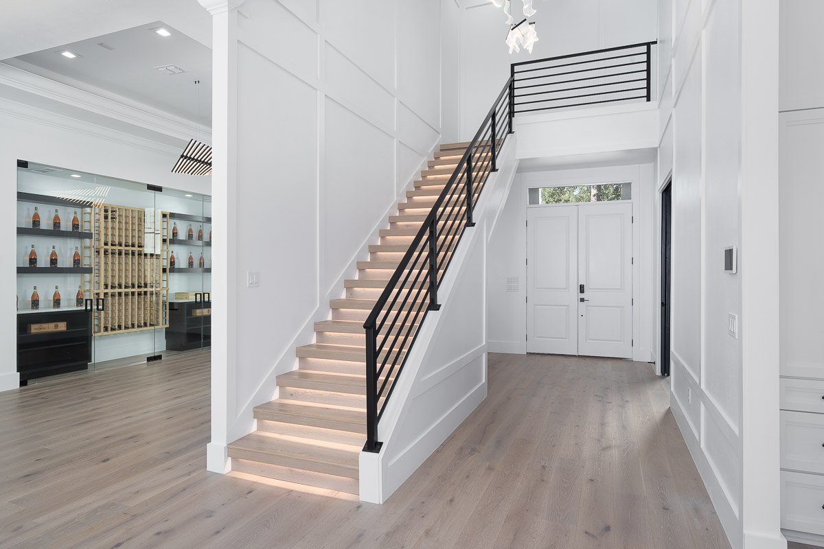 Professional flambient real estate photography of a luxury Seattle entryway with an illuminated staircase, black railings, light wood floors, and custom wine storage.