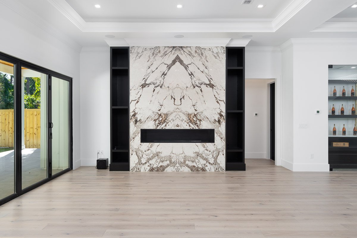 Professional flambient real estate photography of a luxury Seattle living room with a dramatic bookmatched stone fireplace, built-in shelving, large sliding glass doors, and light wood floors.