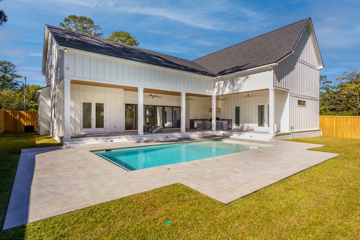 Professional real estate photography of a luxury modern farmhouse-style home with a covered outdoor living area, swimming pool, fenced backyard, and spacious patio.