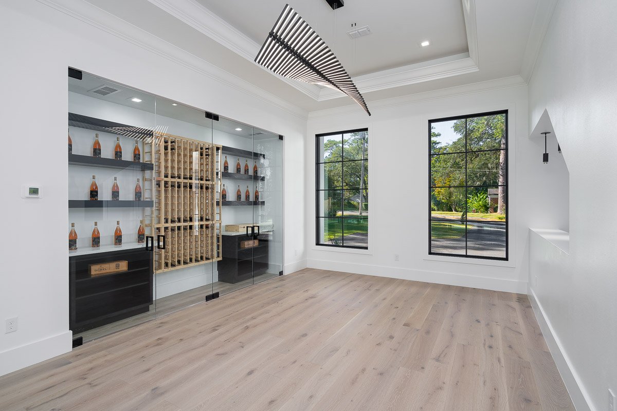 Flambient real estate photography of a bright luxury wine room with a glass-enclosed wine display, tall black-framed windows, white walls, and light wood flooring.