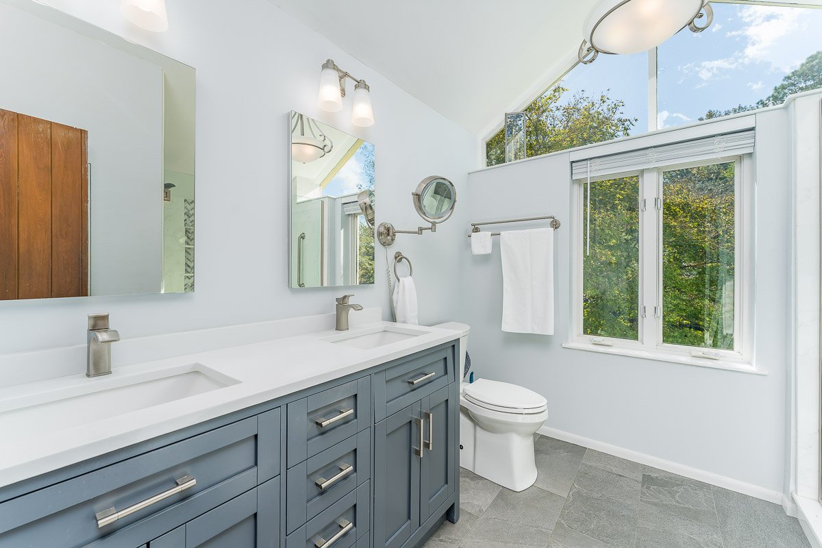 Professional flambient real estate photography of a bright, updated bathroom with blue double vanity, vaulted ceiling, large windows, and clean modern finishes.