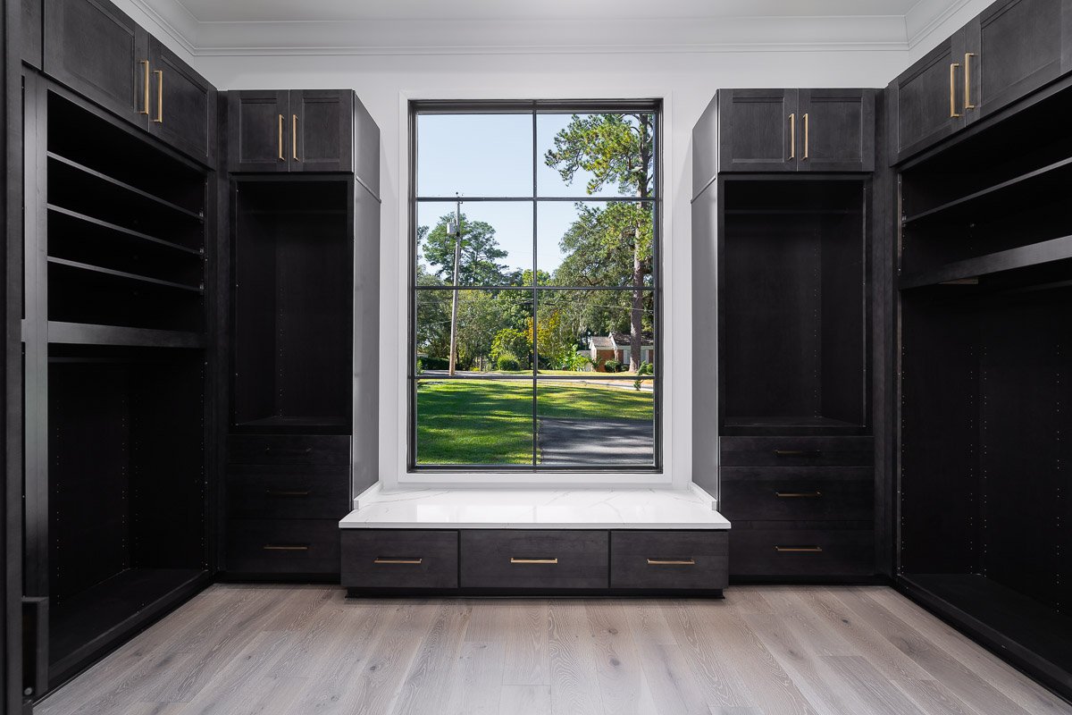 Professional flambient real estate photography of a luxury Seattle walk-in closet with custom dark cabinetry, gold hardware, built-in drawers, a window seat, and abundant natural light.