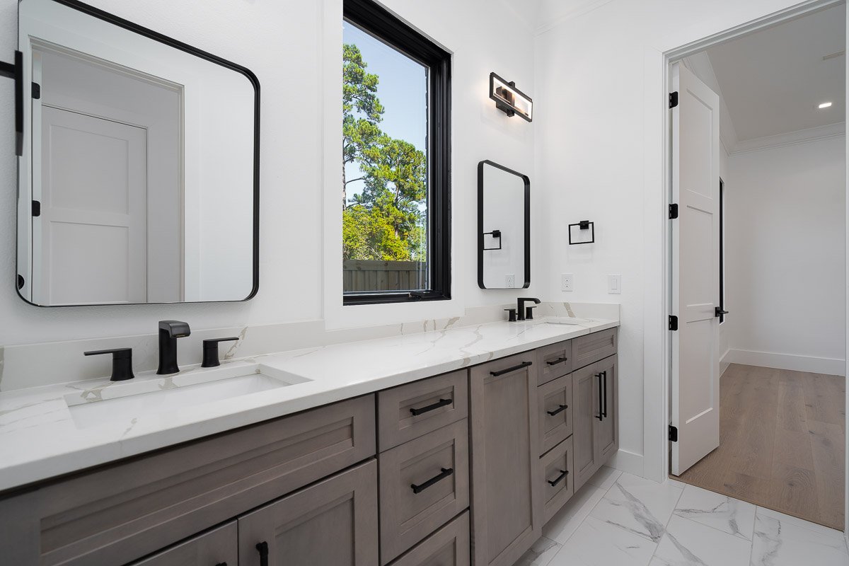 Professional real estate photography of a modern bathroom with a double vanity, quartz countertop, black fixtures, large mirrors, tall window, and bright flambient lighting.