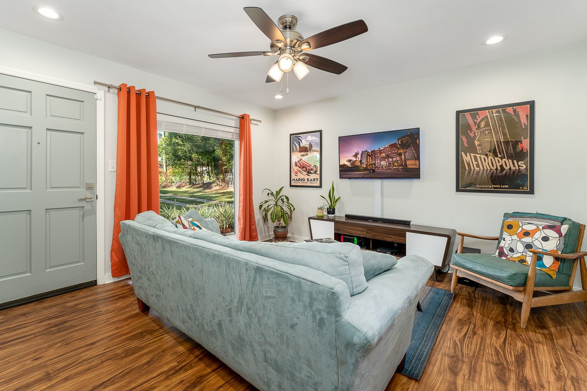 Professional real estate photography of a cozy living room with wood floors, a blue sofa, orange curtains, wall-mounted TV, colorful artwork, and bright flambient lighting.