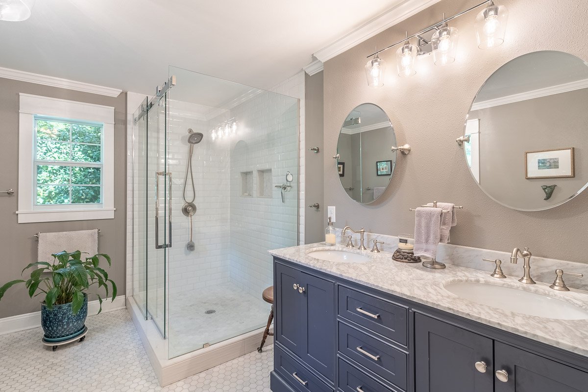 Professional real estate photography of a bright Seattle bathroom with a navy double vanity, marble countertop, glass walk-in shower, dual mirrors, and classic finishes.