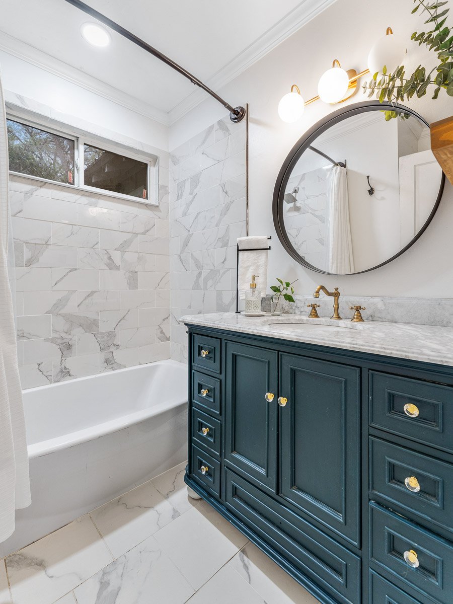 Professional real estate photography of a stylish Seattle bathroom with a deep teal vanity, marble tile, brass fixtures, round mirror, and bright modern finishes.
