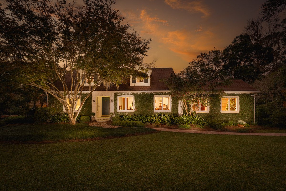 Professional twilight real estate photography of a charming ivy-covered home with warm interior lighting, mature trees, lush landscaping, and a dramatic sunset sky.
