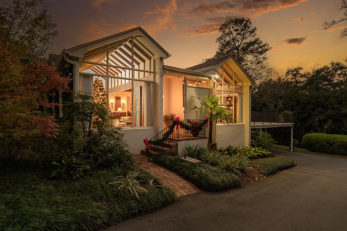 Professional twilight real estate photography of a distinctive modern home with floor-to-ceiling windows, warm interior lighting, lush landscaping, and a dramatic sunset sky.