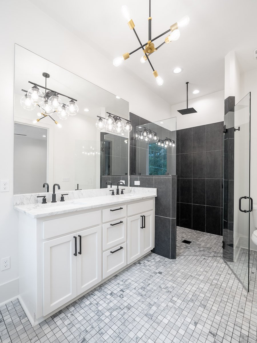 Professional real estate photography of a luxury Seattle bathroom with a double vanity, marble countertop, glass walk-in shower, dark tile, and modern black-and-gold lighting.