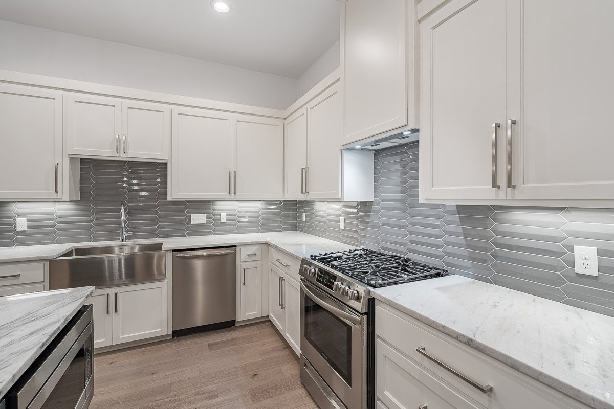 Professional real estate photography of a modern Seattle kitchen with white cabinetry, stainless steel appliances, farmhouse sink, marble countertops, and a geometric gray tile backsplash.