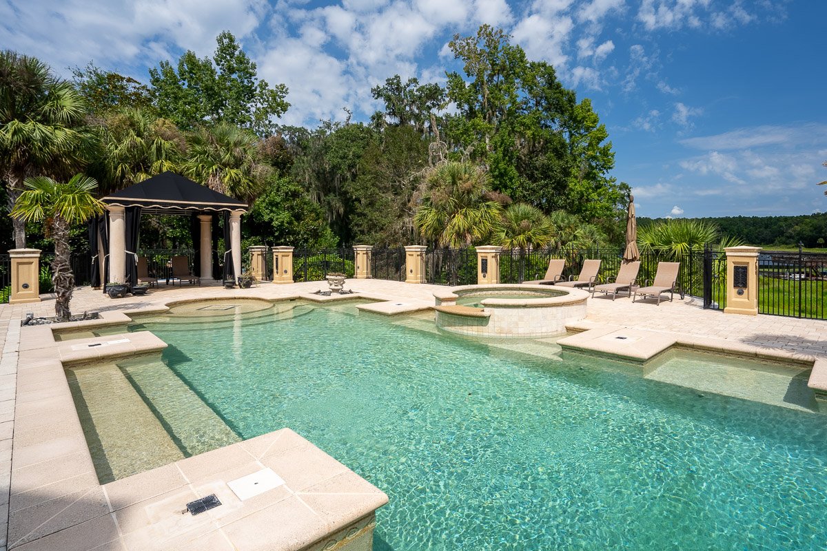 Professional real estate photography of a luxury backyard pool with a raised spa, tropical landscaping, elegant stonework, and resort-style outdoor living.