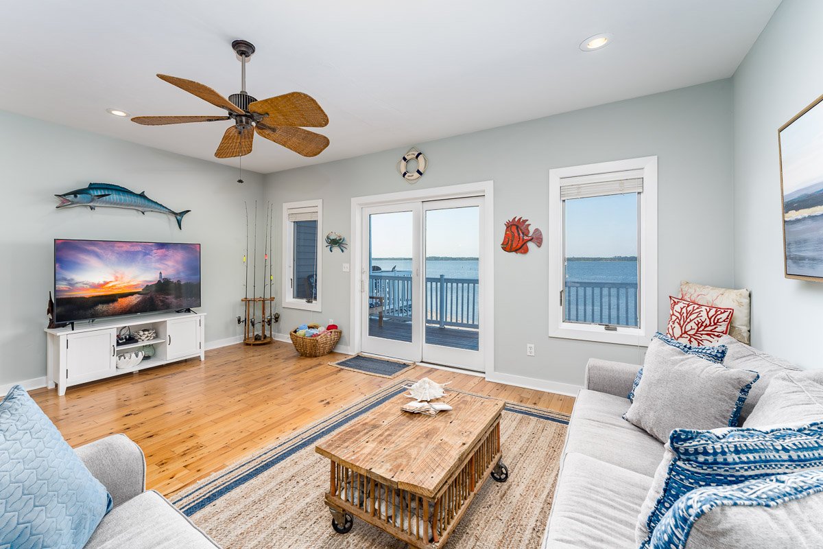 Flambient real estate photo of a bright coastal living room with water views, sliding glass doors to a deck, light wood floors, and beach-inspired decor.