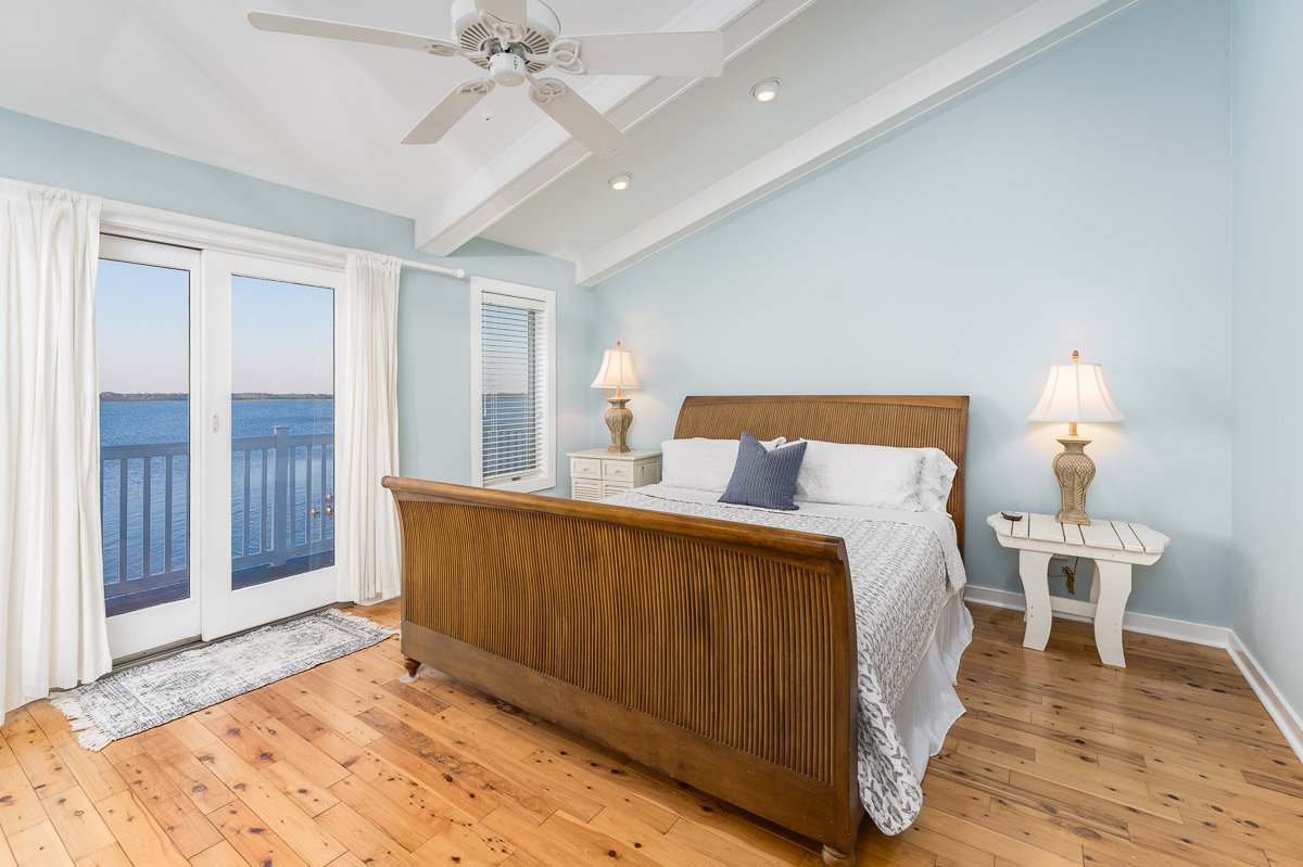 Professional real estate photography of a bright waterfront bedroom with vaulted ceilings, wood floors, French doors to a private deck, and expansive water views.
