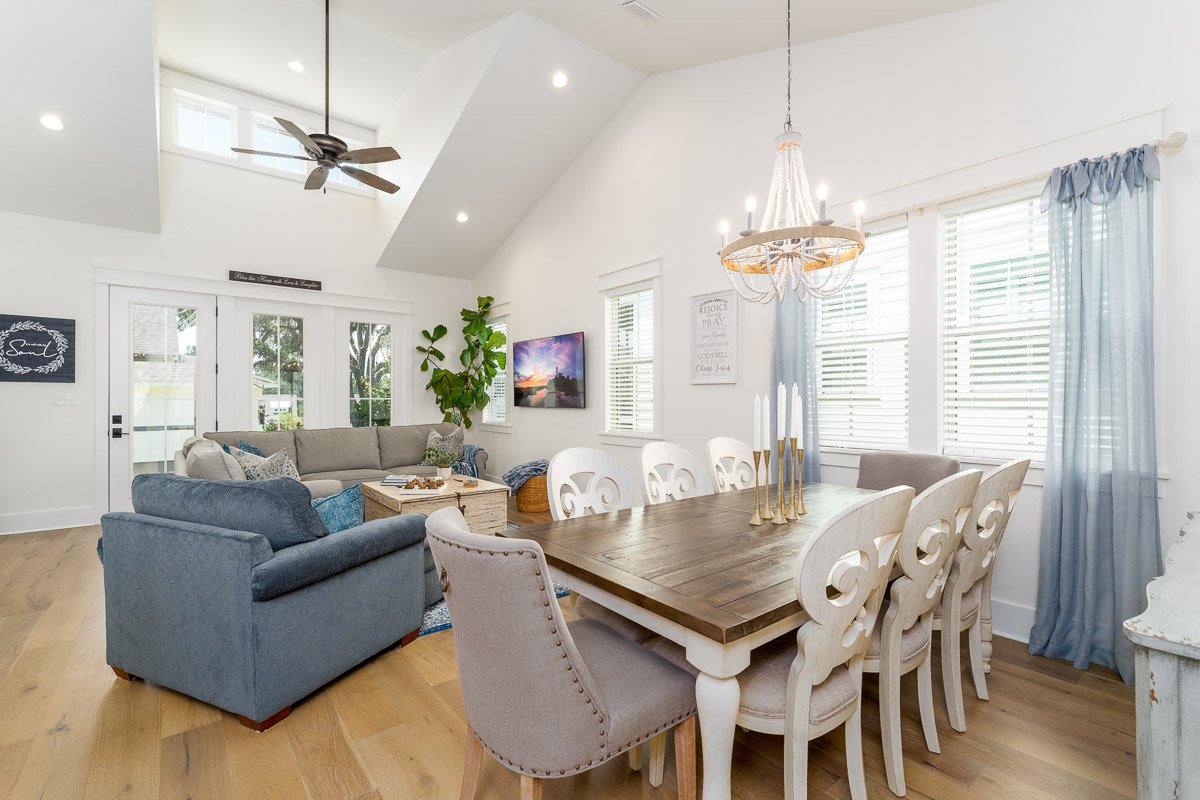 Professional real estate photography of a bright open-concept dining and living area with vaulted ceilings, large windows, elegant chandelier, blue seating, and a spacious coastal-inspired interior.