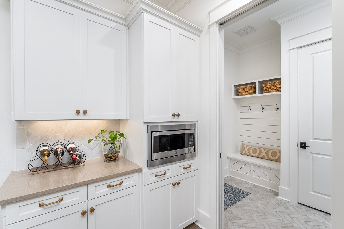 Professional real estate photography of a bright white butler’s pantry with custom cabinetry, built-in microwave, stone countertops, and an adjoining mudroom with bench seating and herringbone tile flooring.
