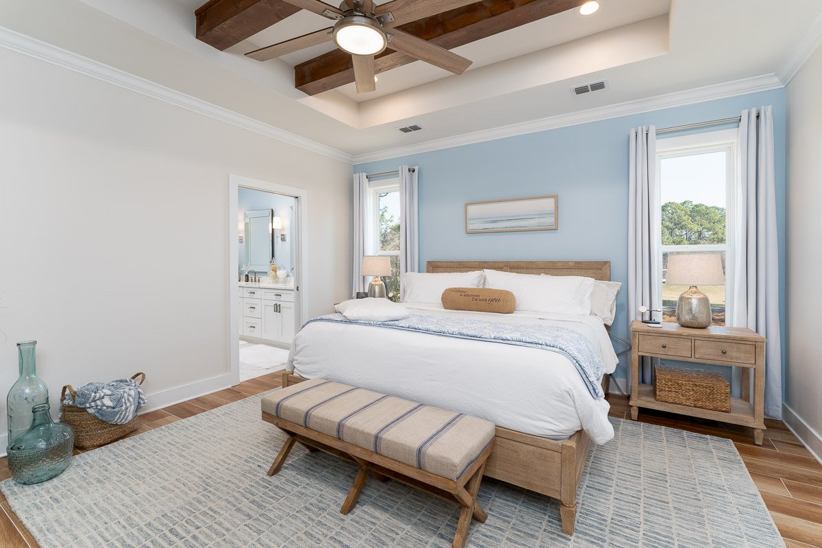 Professional flambient real estate photography of a bright primary bedroom with a tray ceiling, exposed wood beams, blue accent wall, en suite bathroom, and elegant coastal-inspired staging.