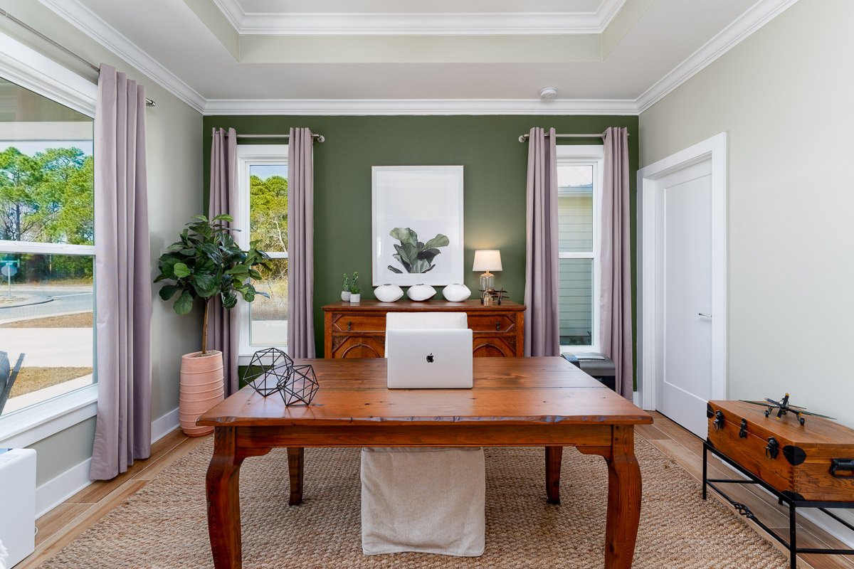 Professional flambient real estate photography of a bright home office with a large wood desk, green accent wall, tall windows, tray ceiling, and stylish contemporary staging.