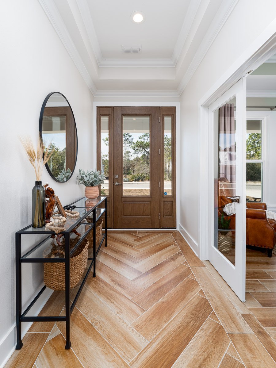 Professional flambient real estate photography of a bright entryway with a wood front door, herringbone tile flooring, decorative mirror, and stylish modern staging.