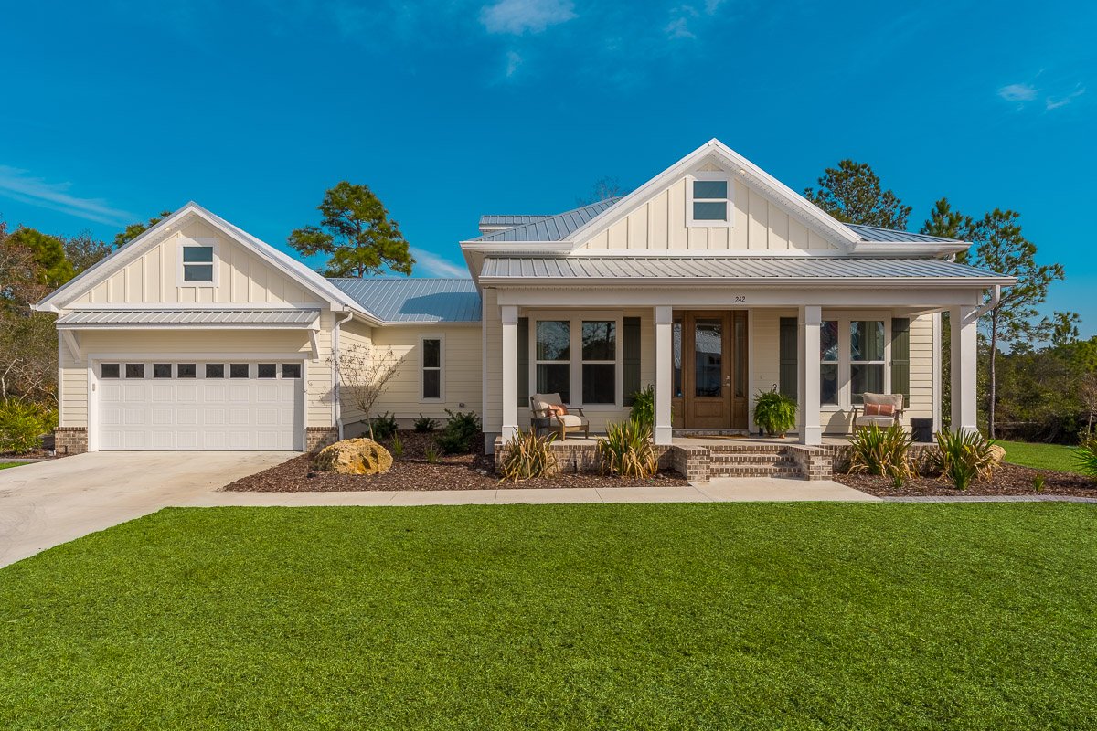 Professional real estate photography of a charming modern farmhouse-style home with a metal roof, covered front porch, attached garage, manicured lawn, and inviting curb appeal.